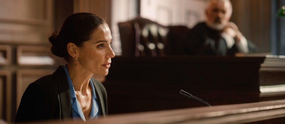 Courtroom scene with a judge sitting on the bench in a blurred background and a woman sitting in the foreground at the witness stand in front of a microphone.