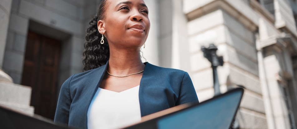 A woman standing in front of a blurred courthouse, looking to the right, holding an open binder.